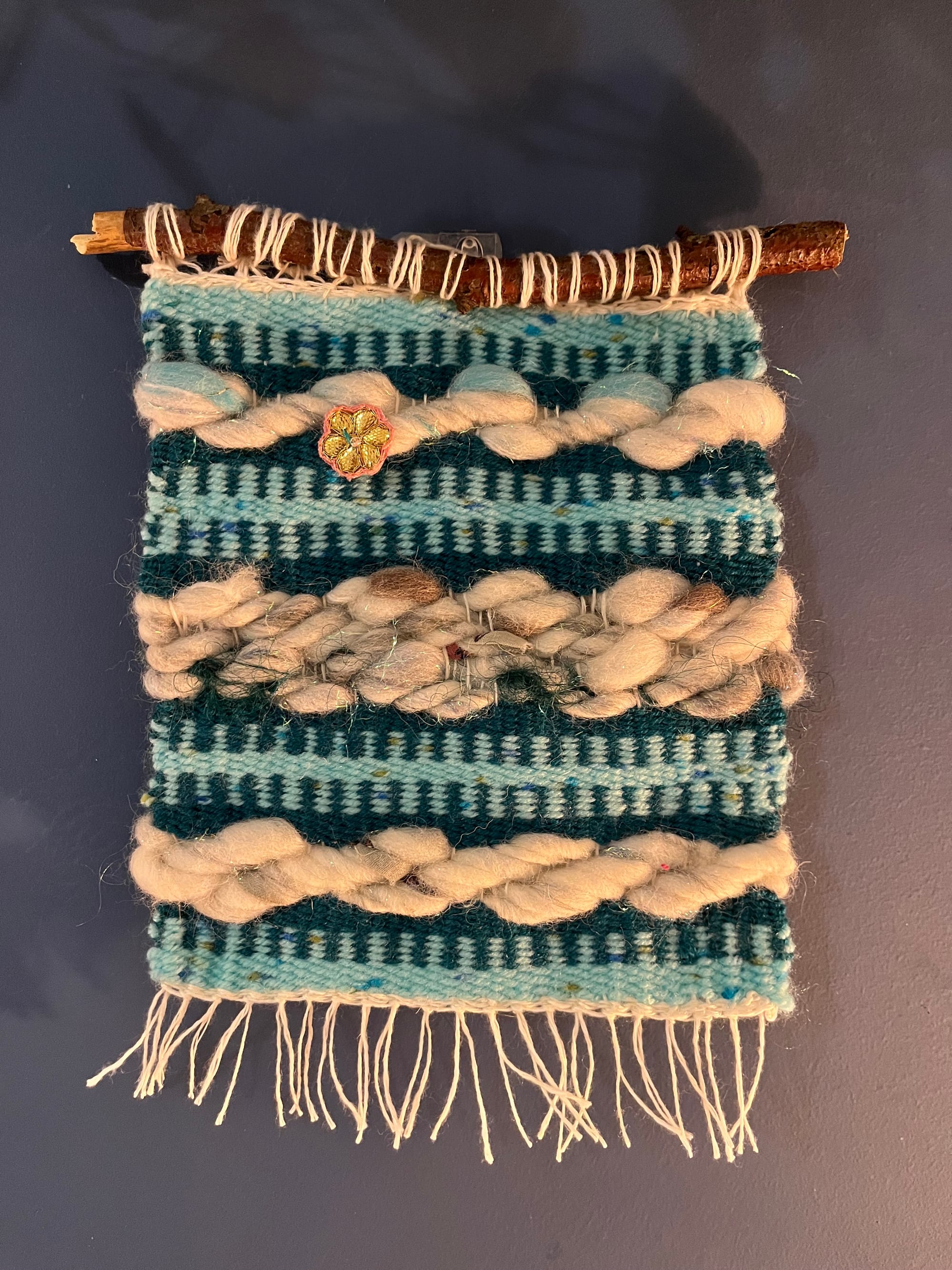 A small tapestry woven from sky blue yarn, dark teal yarn, and grey/white yarn that is thickly spun and pops off the piece. It hangs from a dowel made from a slightly curved foraged stick.